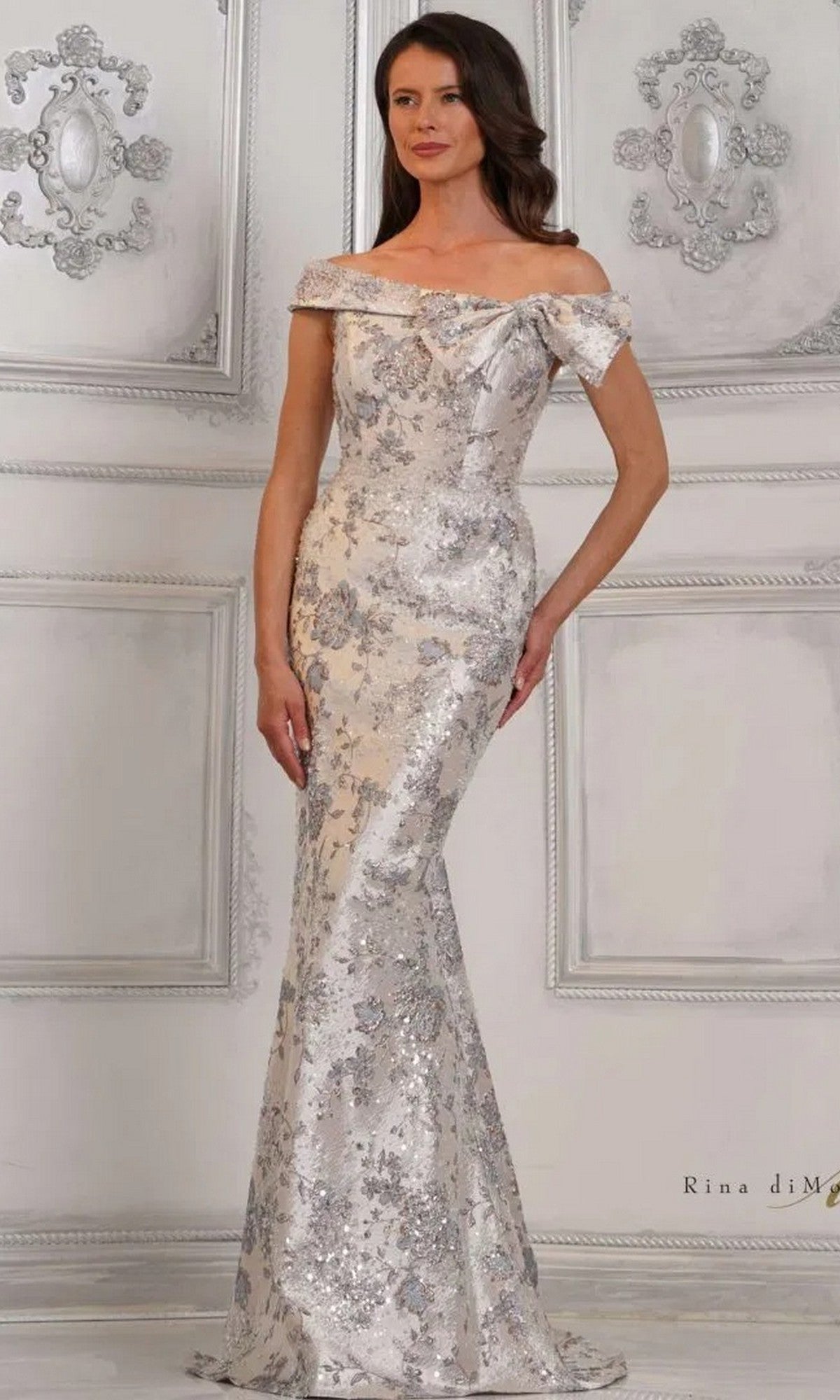 A woman in the Rina Di Montella RD3106 long formal dress, featuring a blue multi-color design and off-the-shoulder style, stands against an ornate white-paneled wall.